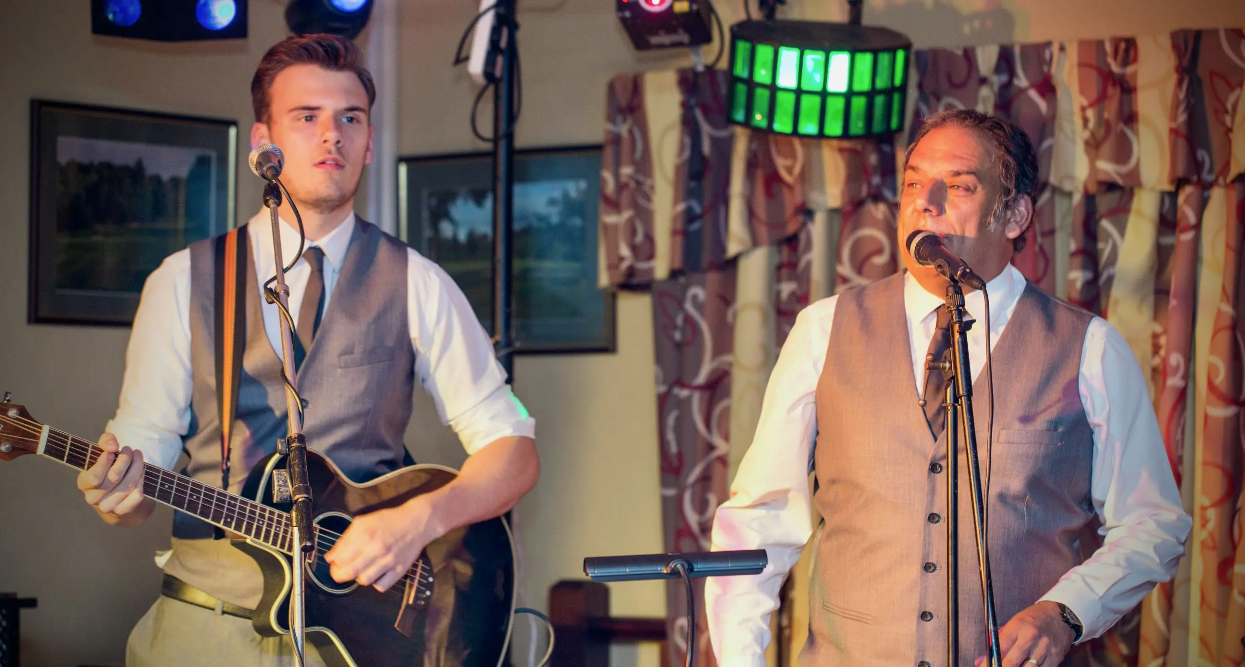 Acoustic Duo Eastbound and Down on stage singing into microphones dressed in white shirts, dark tie and grey waistcoats Jack is playing a guitar